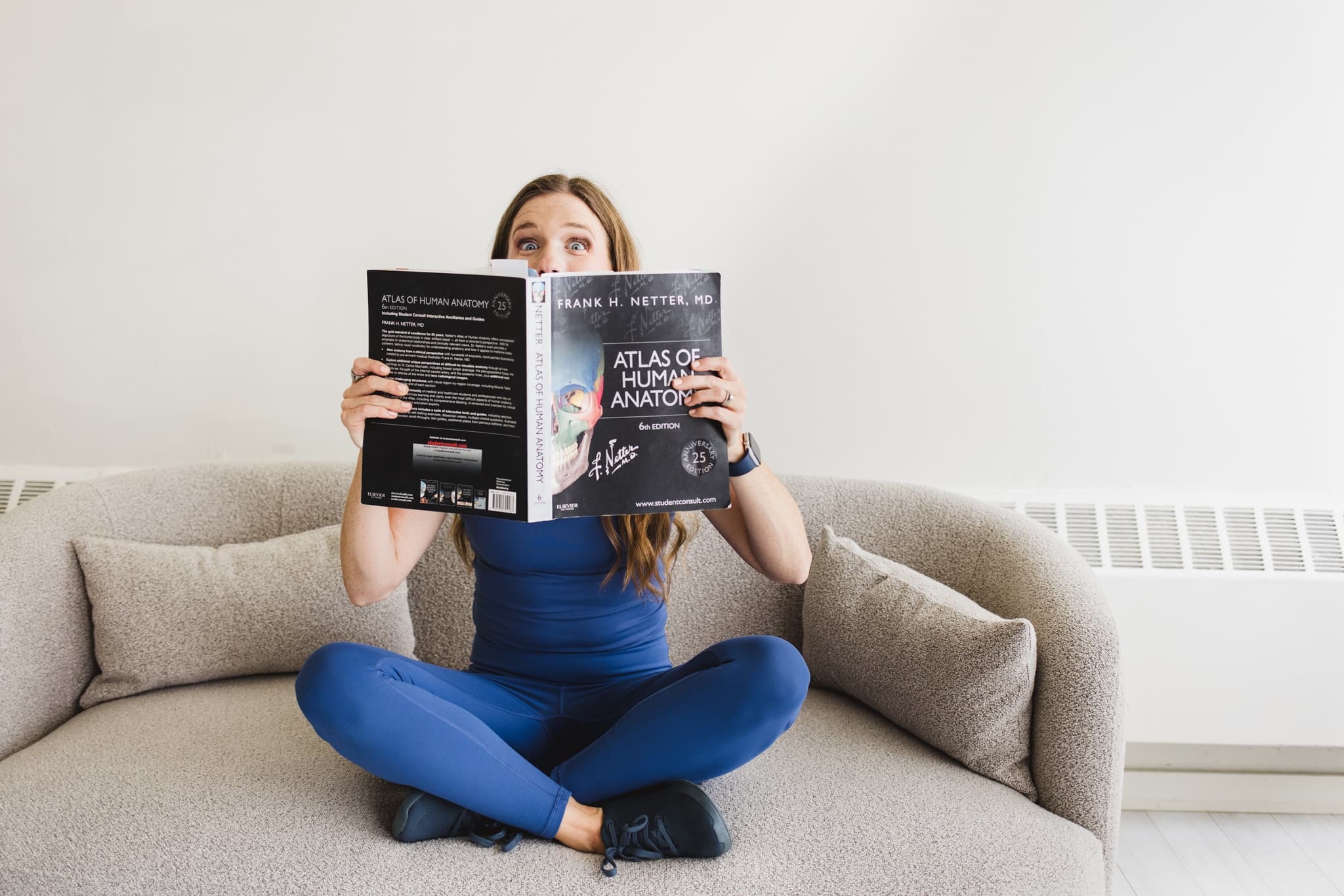 Pelvic floor physical therapist sitting cross-legged on a couch while holding an anatomy book and looking over top of the book with excitement in her eyes.