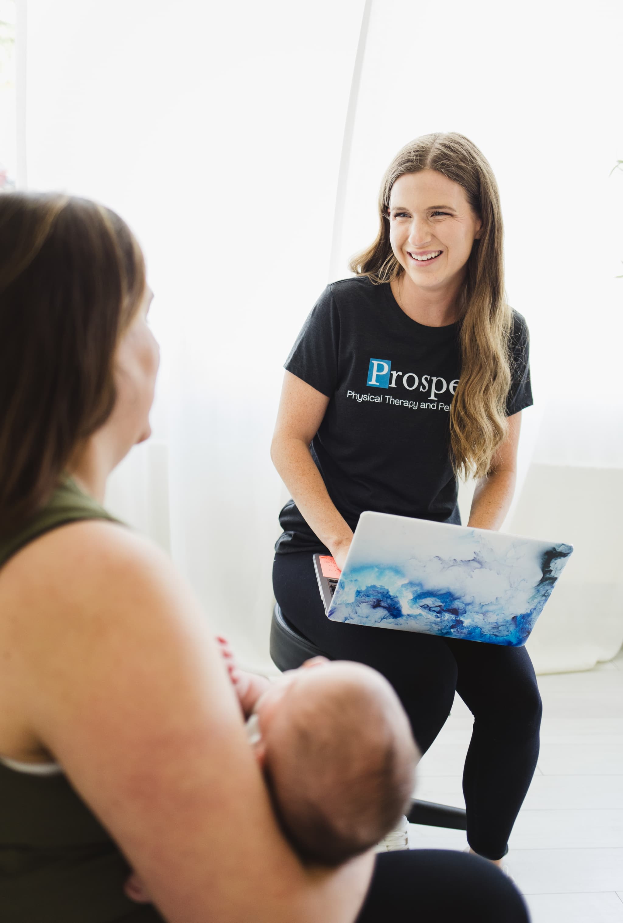 Pelvic floor physical therapist typing on her laptop while having a conversation with a recently postpartum mom coming in for her first visit after having her baby.