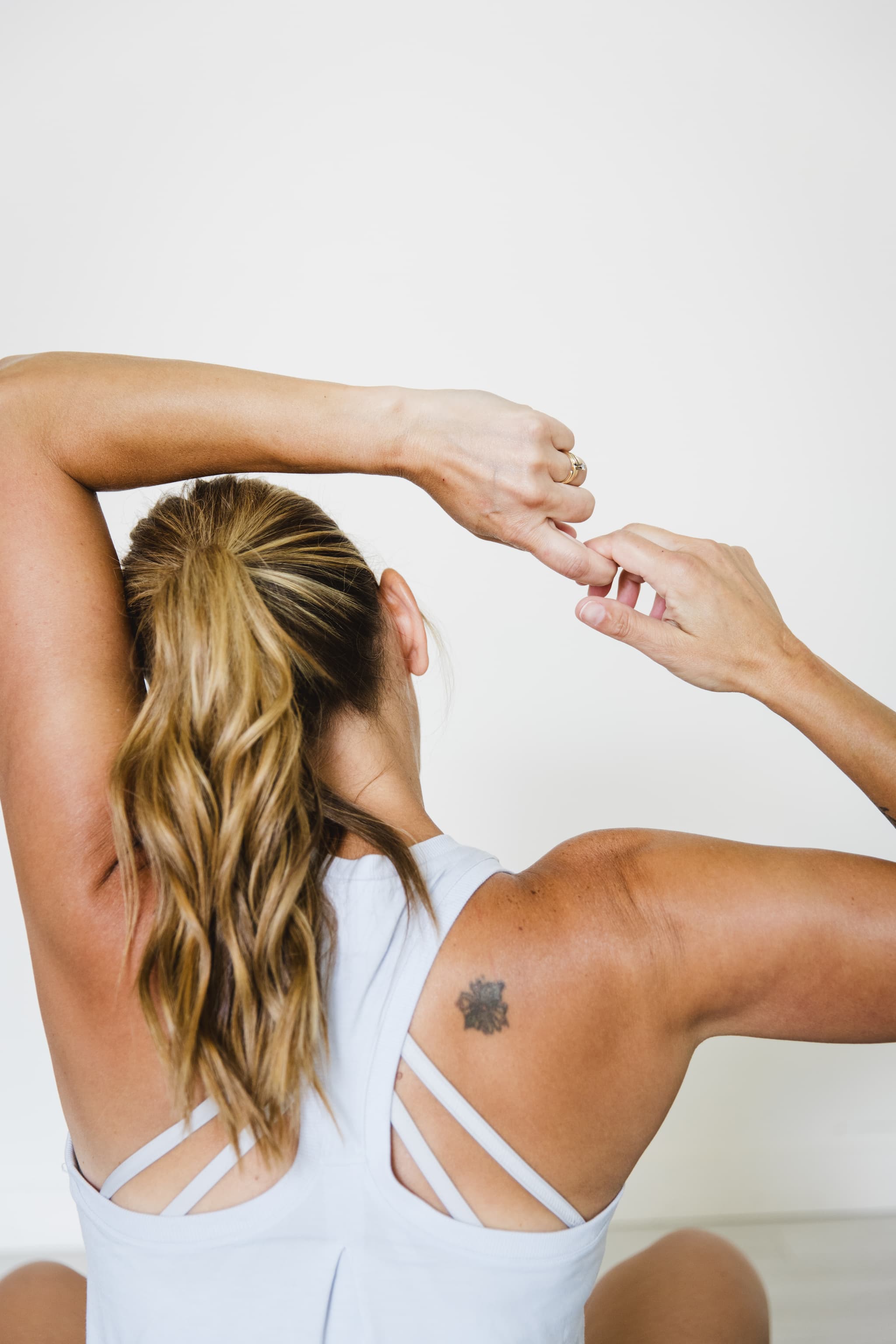A woman is facing away in a seated position reaching one hand over her head to interlock fingers to stretch her neck.